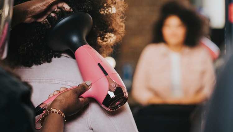 A hair stylist using a pink ghd hairdryer on a person with curly brown afro hair