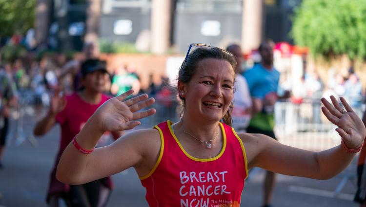 A woman with brown hair is smiling and waving during her race. She is wearing a pink Breast Cancer Now running vest.